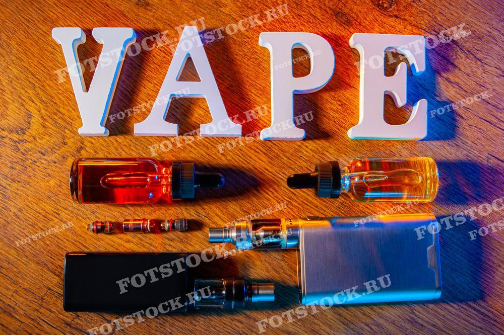 На фотографии изображены буквы, составляющие слово "VAPE", и различные аксессуары для вейпинга, такие как электронные устройства и флаконы с жидкостью. Все элементы расположены на деревянной поверхности.